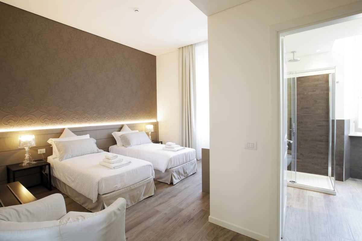 A modern hotel room with two beds, a sitting area, and a glass shower enclosure.