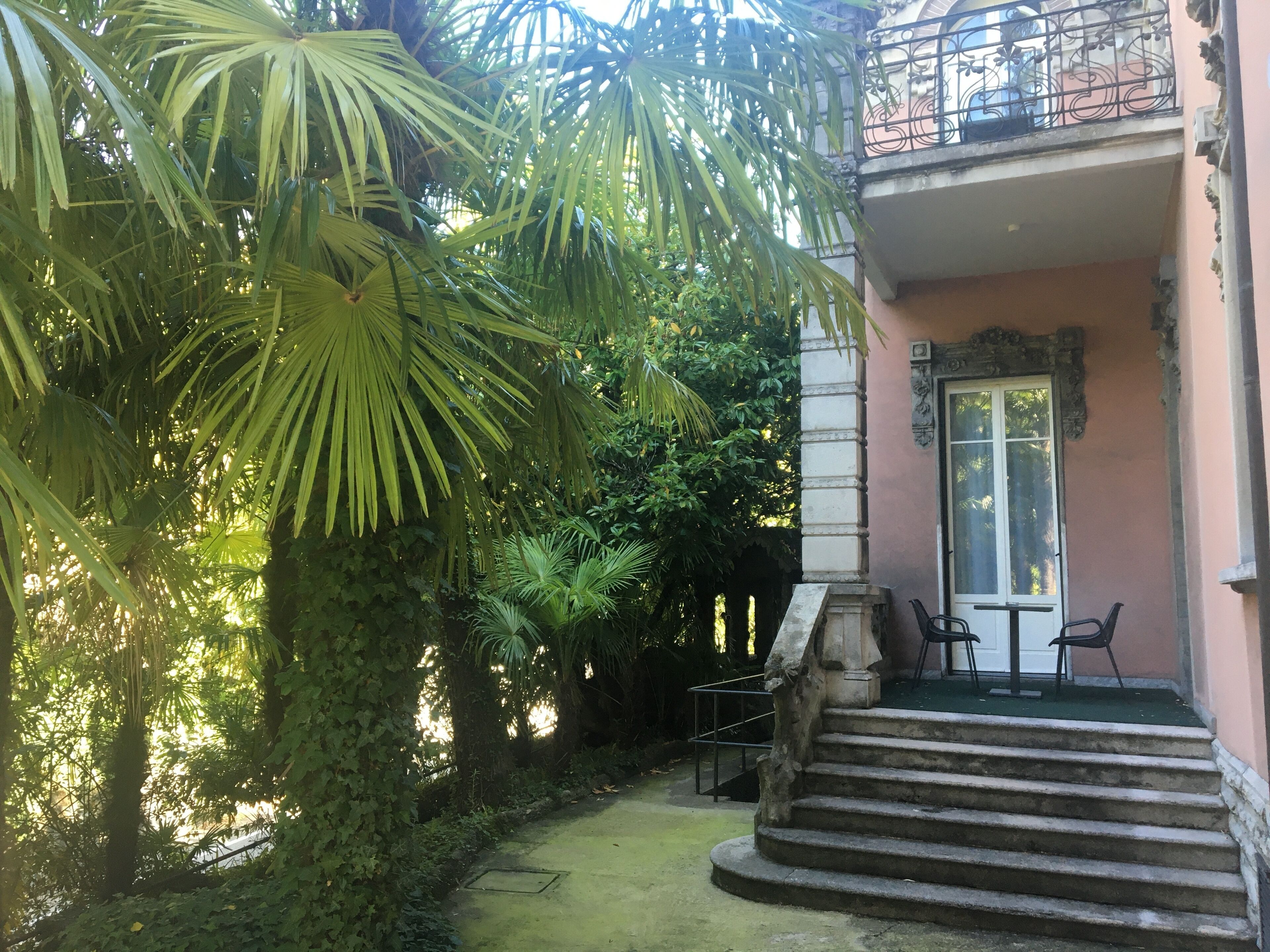 Un edificio con balcón, una escalera que lleva a la entrada y un jardín con diversas plantas.