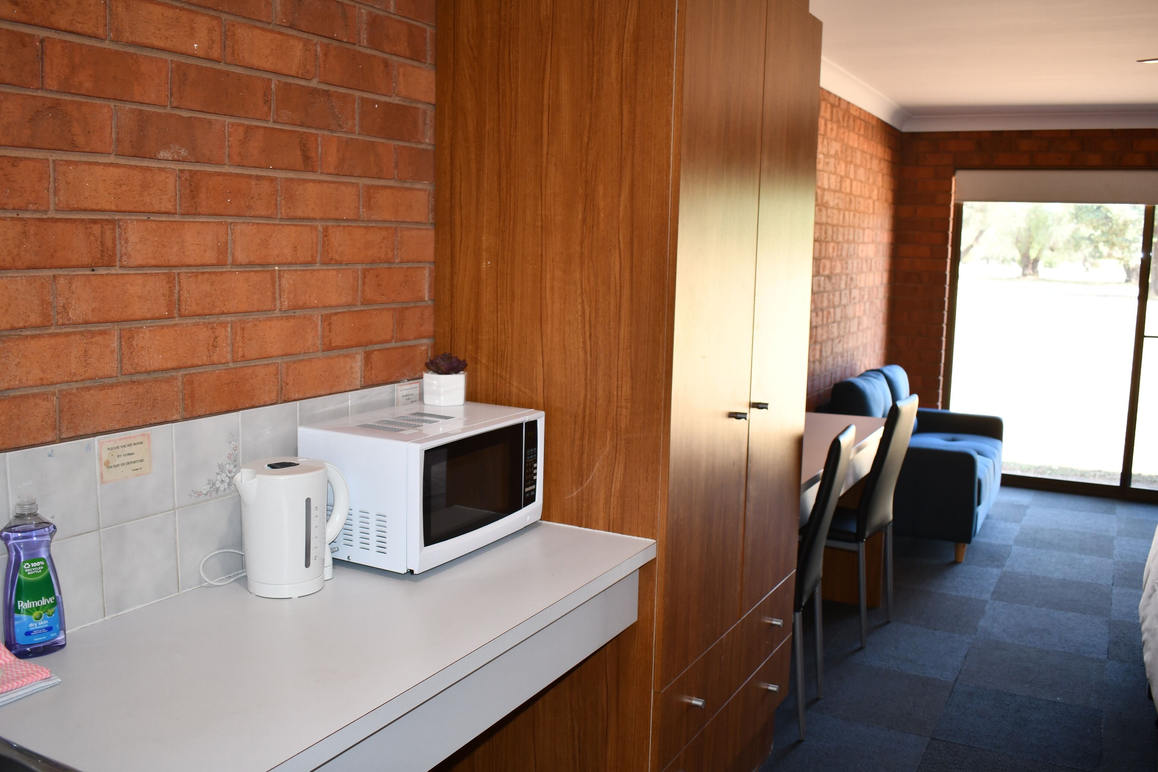 A kitchen with a microwave, kettle, and dish soap on the counter. There is a brick wall and a wooden cabinet.