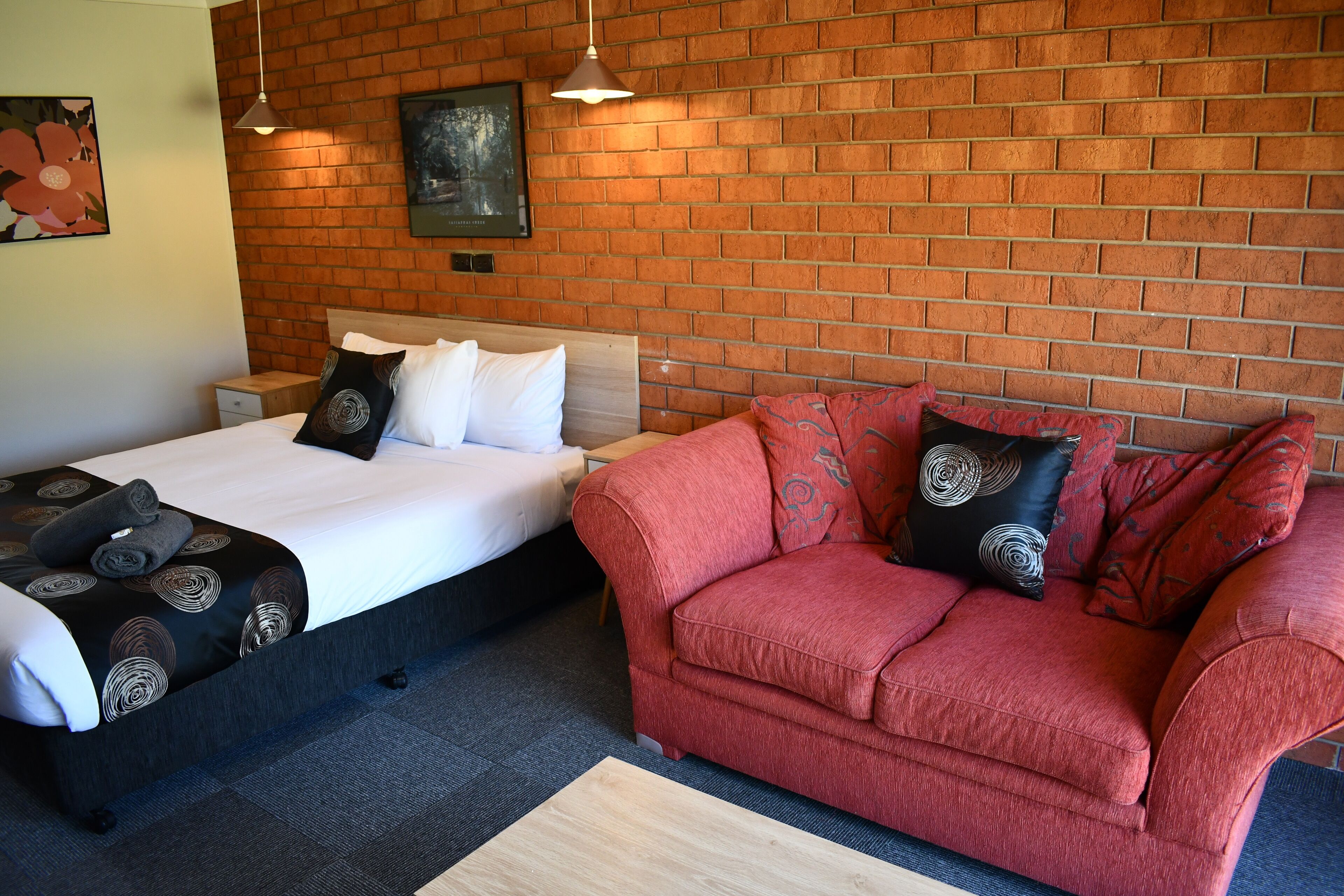 Ein Hotelzimmer mit Backsteinwand, einem Bett mit gemusterten Betttextilien, einem roten Sofa mit dekorativen Kissen und einem kleinen Holfnachtkästchen.