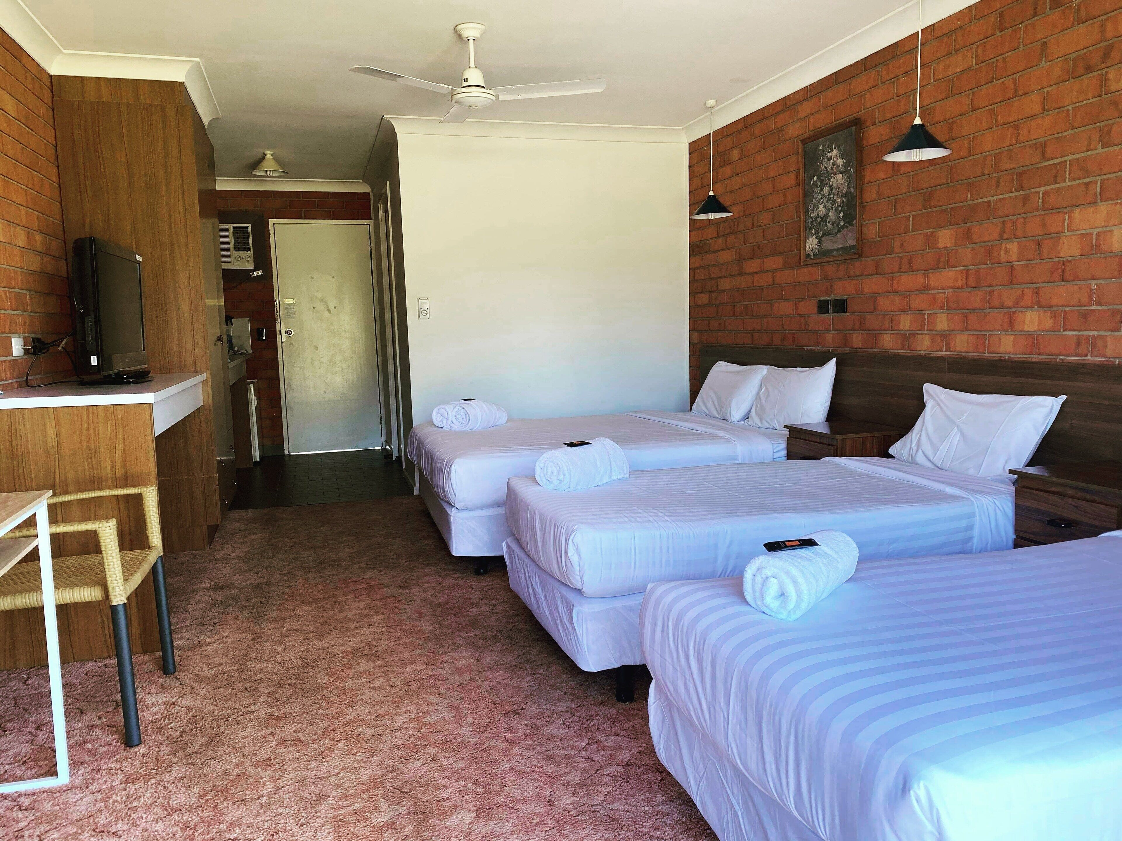 Habitación de hotel con dos camas, un escritorio, un televisor, un ventilador de techo y una pared de ladrillo.