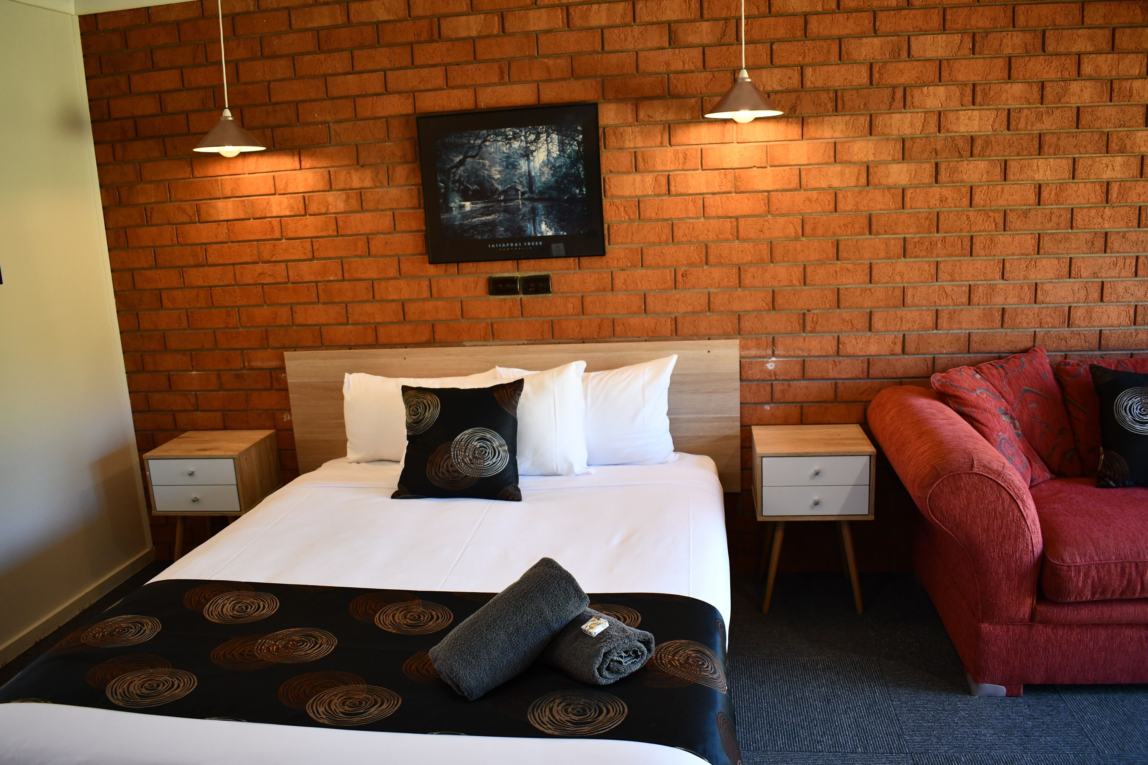 A hotel room with a brick wall, a bed with white bedding, two bedside tables, a red armchair, and a framed picture on the wall.
