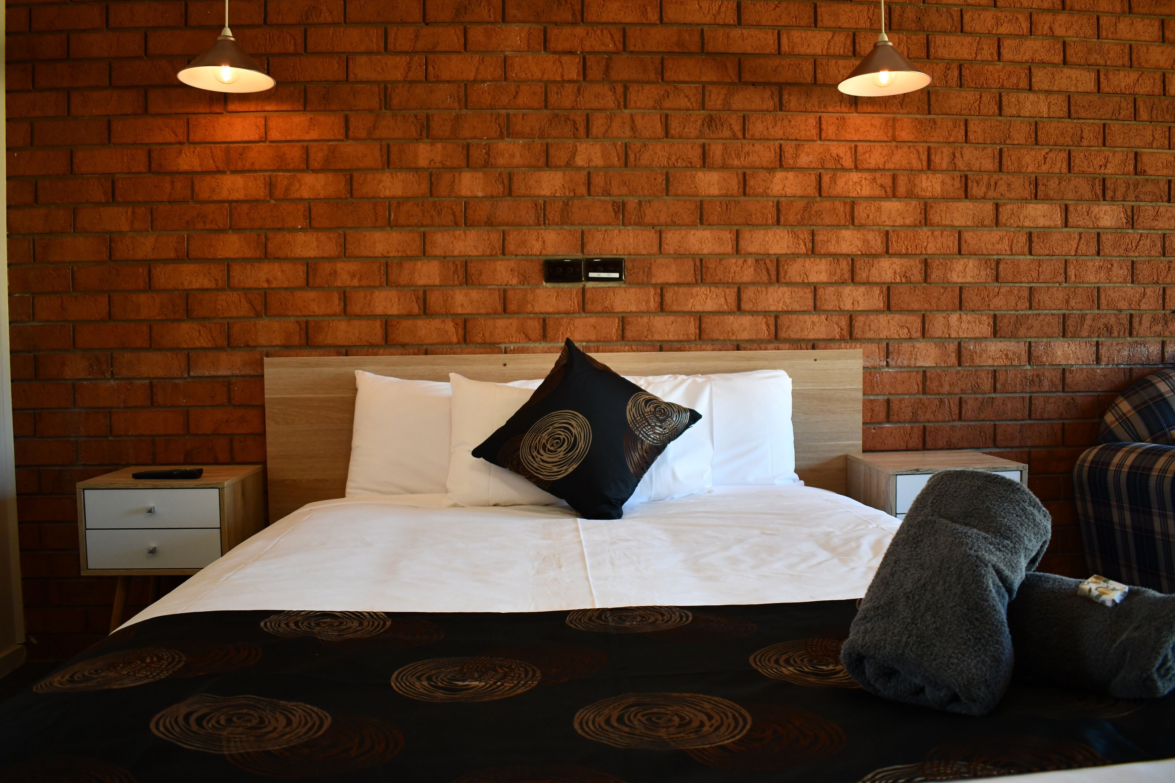 A hotel room with a brick wall, a wooden headboard, a bed with white bedding and a black pillow, a nightstand, and a lamp.