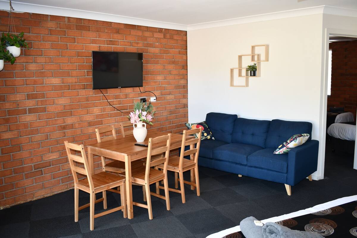 Un salon avec un mur en briques, un téléviseur à écran plat fixé au-dessus d’une table à manger en bois, entourée de chaises, un canapé bleu et une petite plante accrochée au mur.