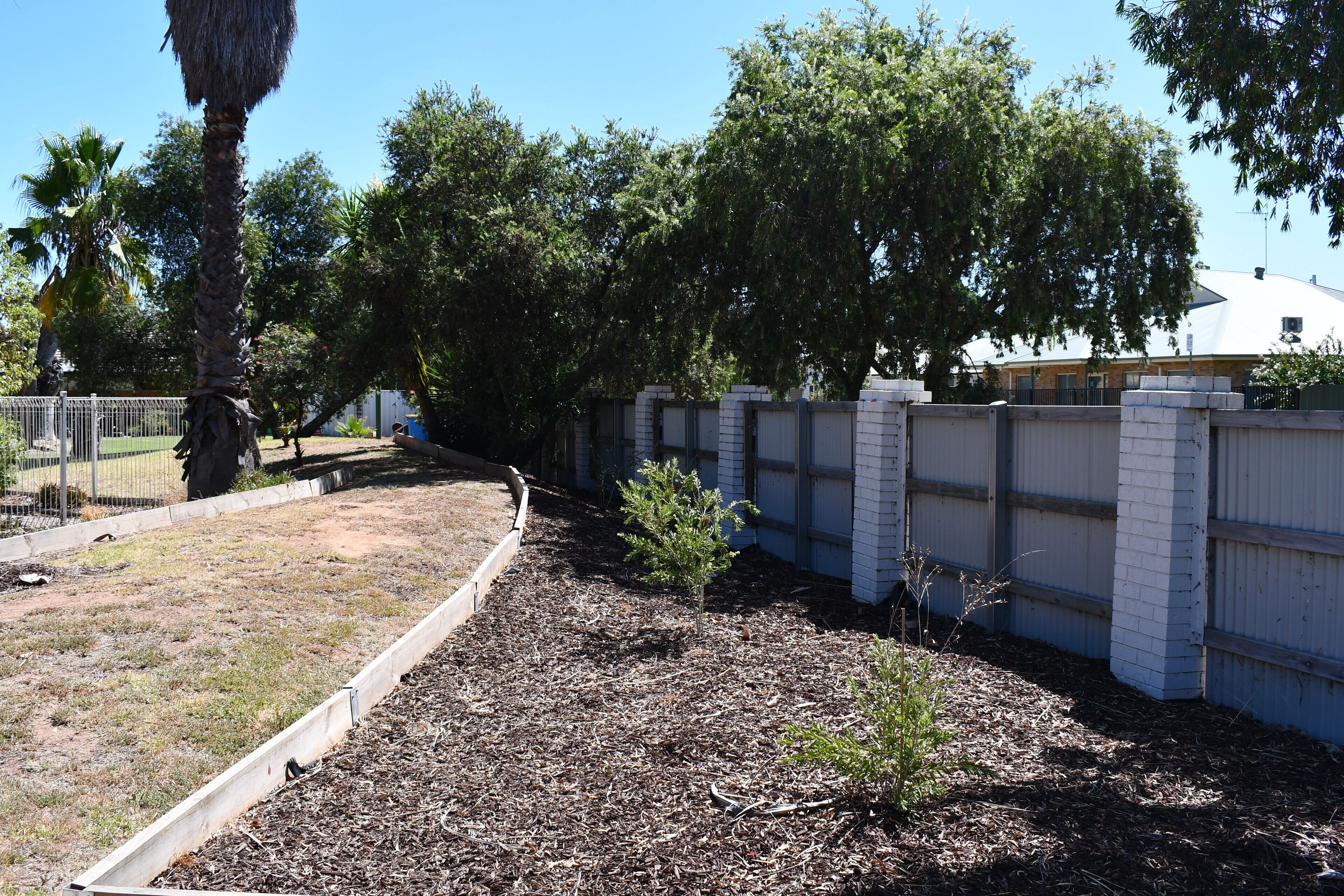 A fenced yard with trees, a small garden bed, and a white building in the background.