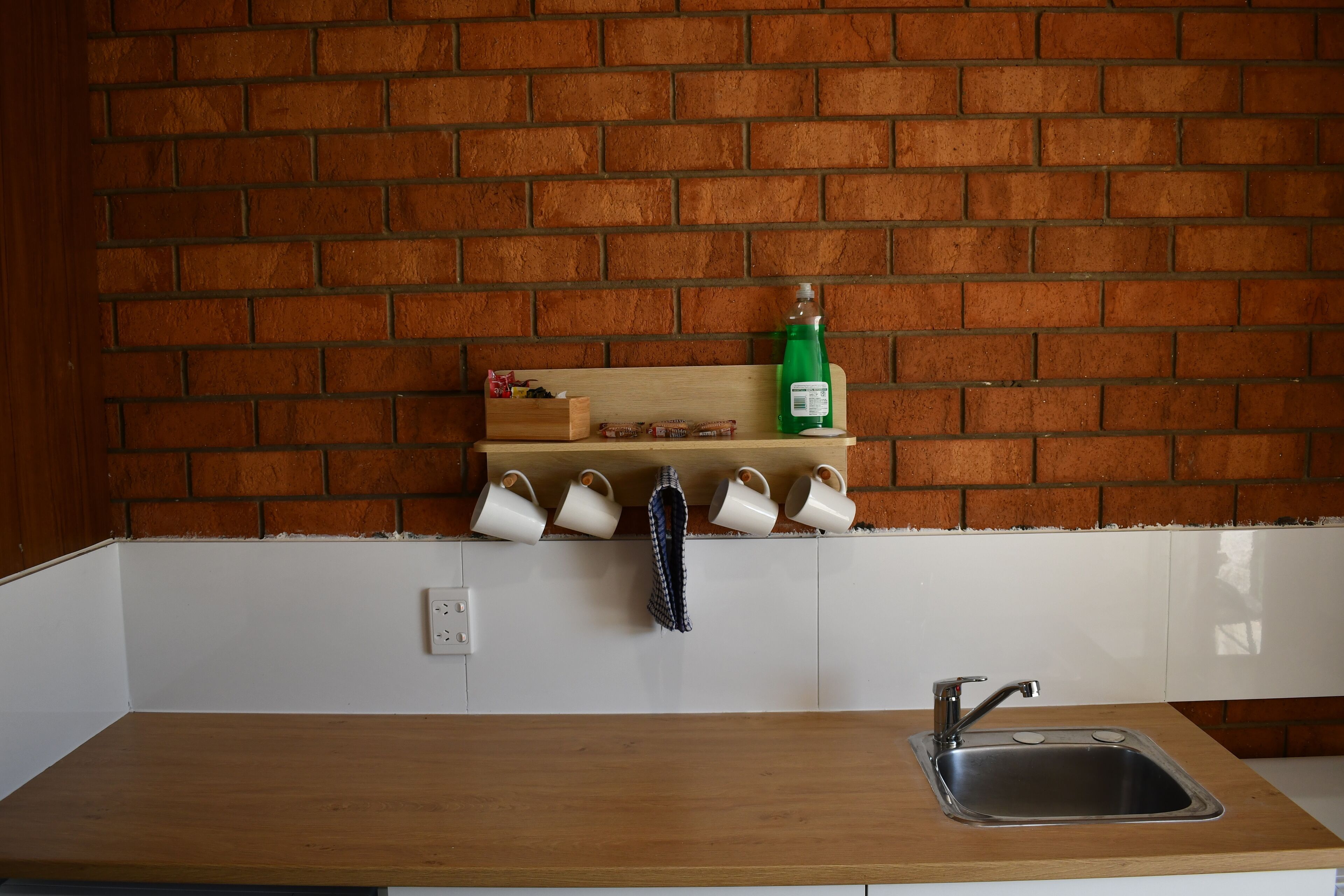Una cocina con pared de ladrillo, encimera de madera, un fregadero y una estantería con tazas y una botella.