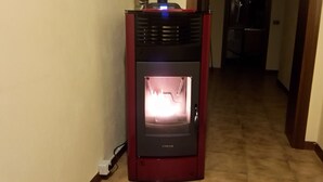 A red wood-burning stove with a visible fire inside, located in a hallway with a tiled floor. - Gessy's (Lucca)