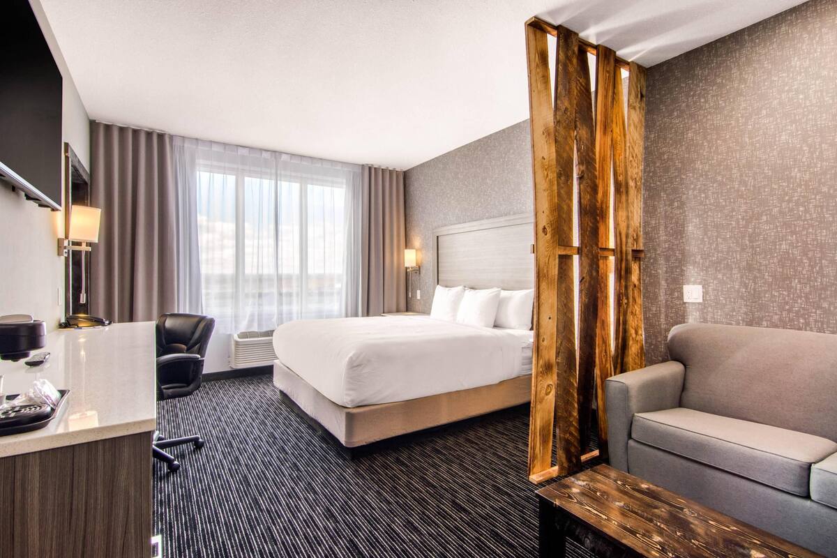 A hotel room with a bed, a desk, a chair, a sofa, and a large window with curtains.