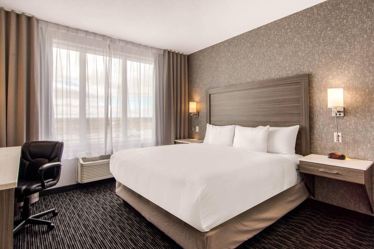 A hotel room with a large bed, a desk, a chair, a nightstand with a lamp, and a view of the outdoors through sheer curtains.