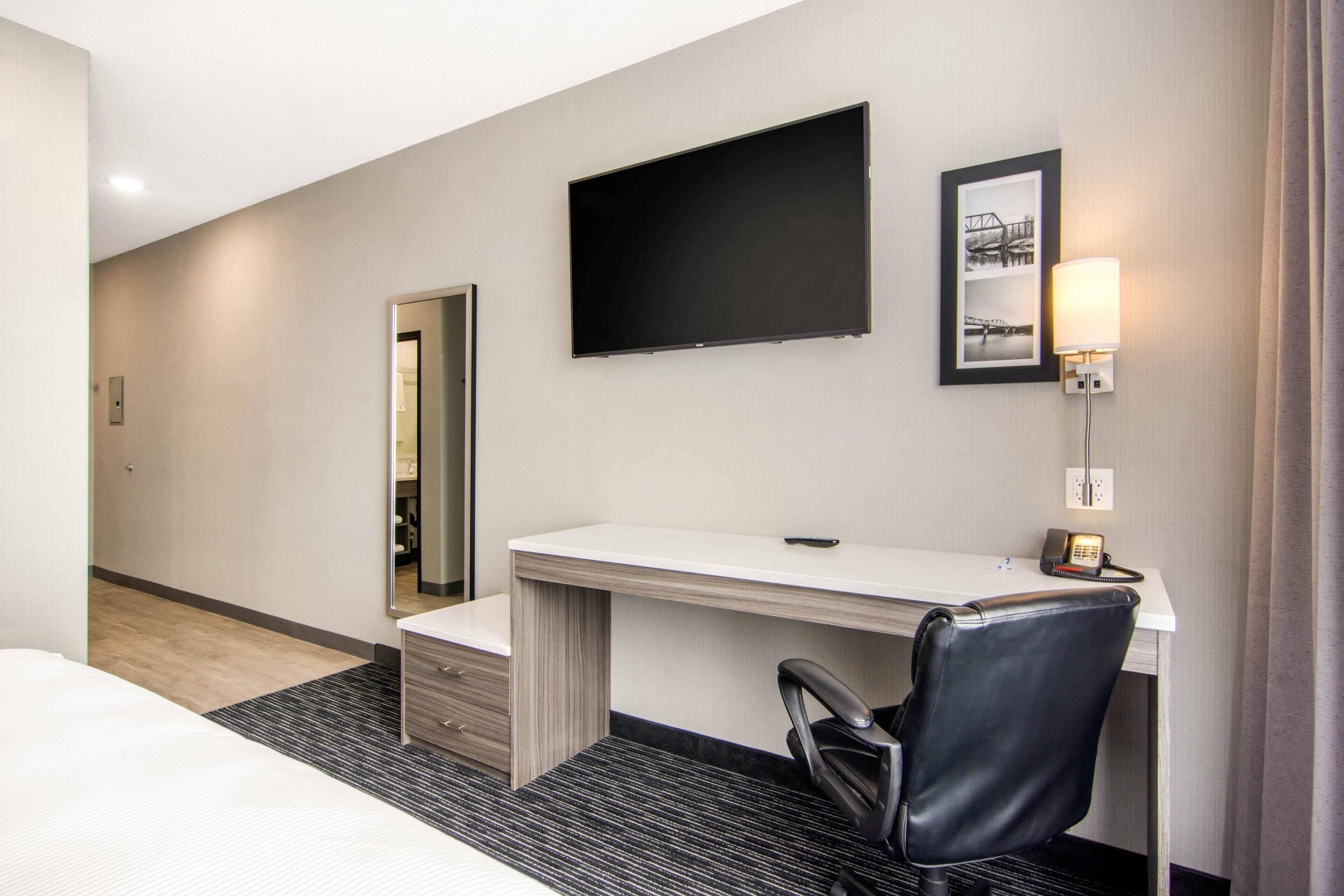 A hotel room with a bed, a desk with a chair, a flat-screen TV, a framed picture, and a bedside lamp.