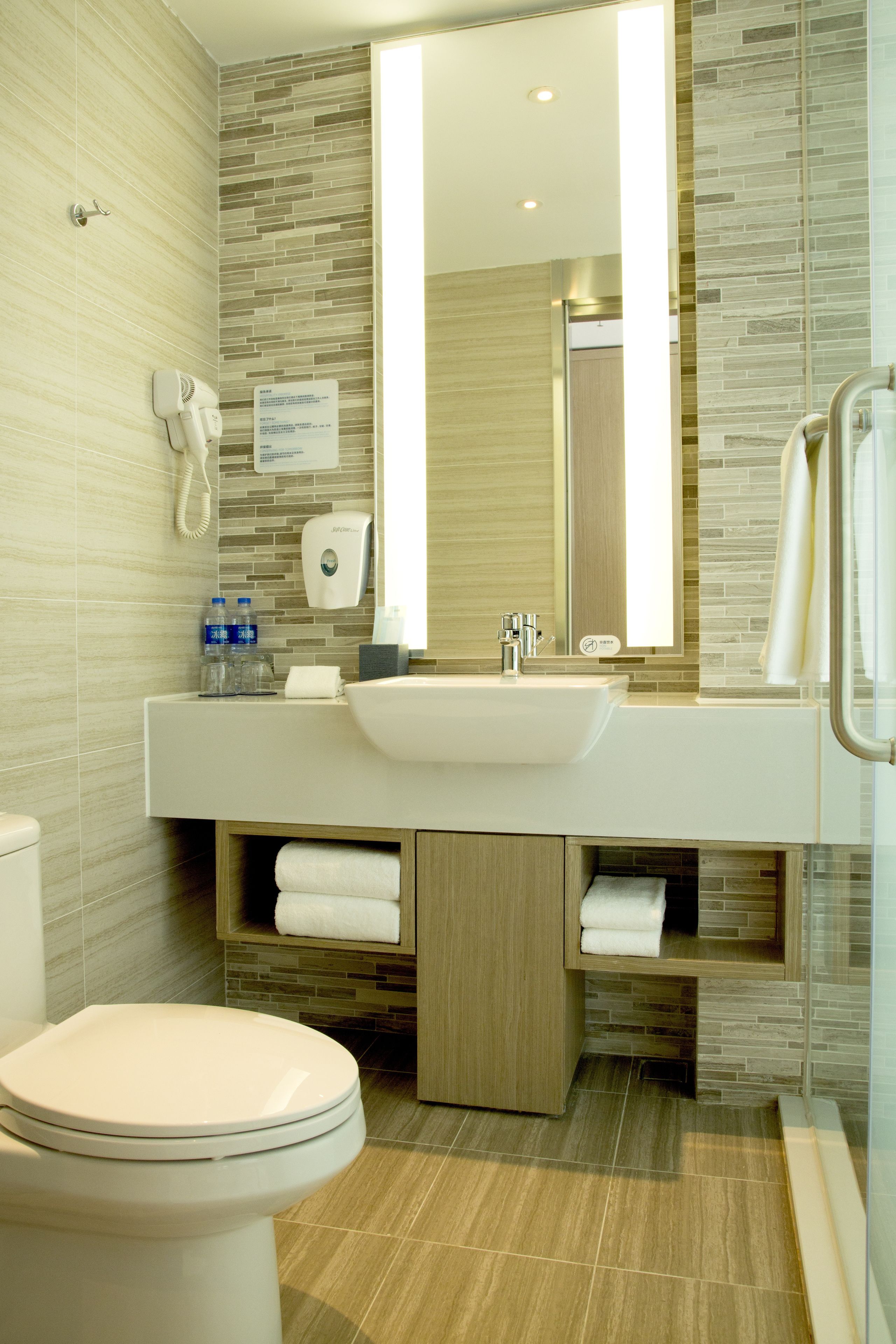A modern bathroom with a large mirror, a sink, and a toilet. There are towels and a hand dryer on the counter.