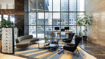A modern lobby with a curved sofa, armchairs, a coffee table, and a small table with two monitors. Large windows offer a view of the cityscape.