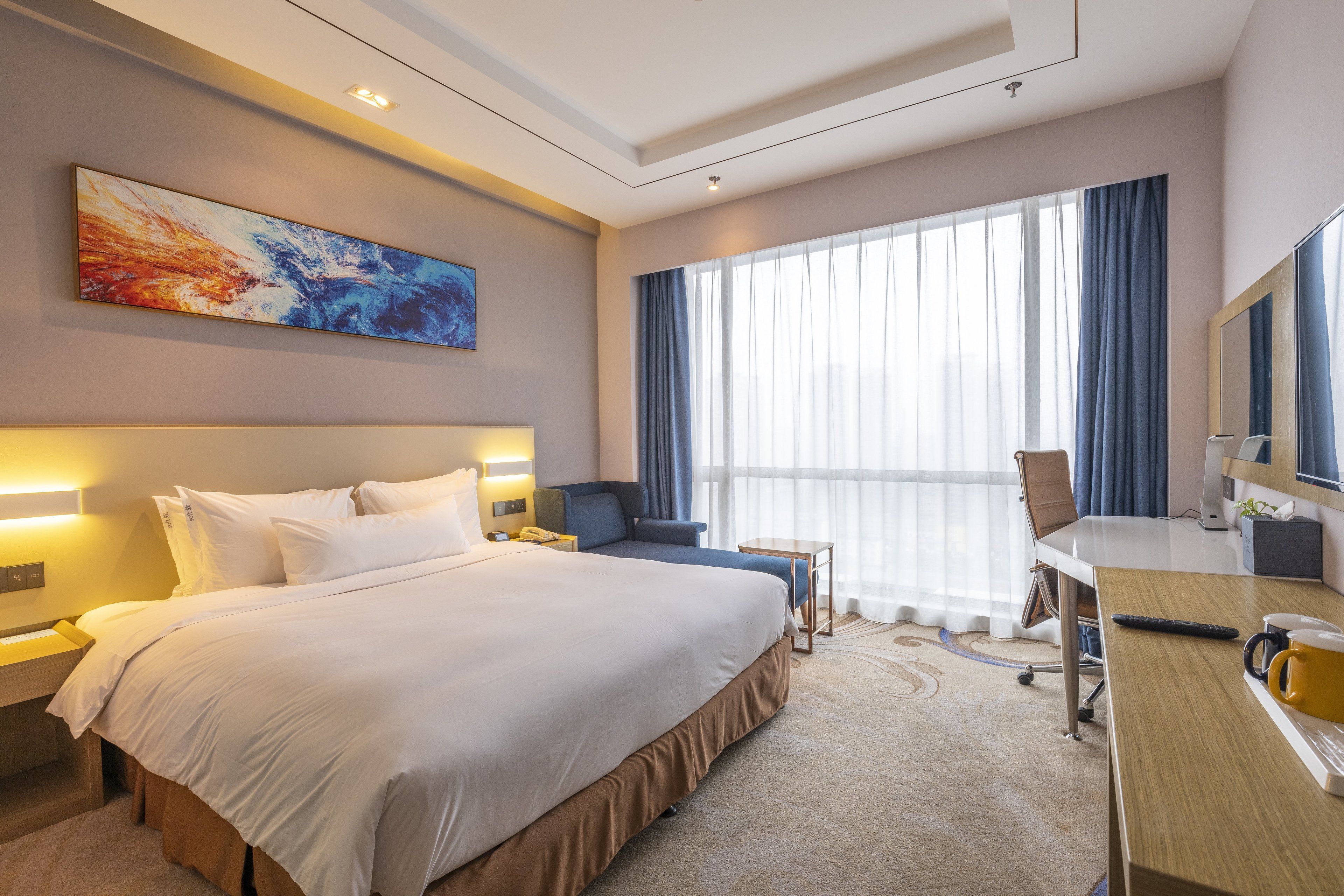 A hotel room with a large bed, a desk, a chair, a desk lamp, a television, and a window with curtains.