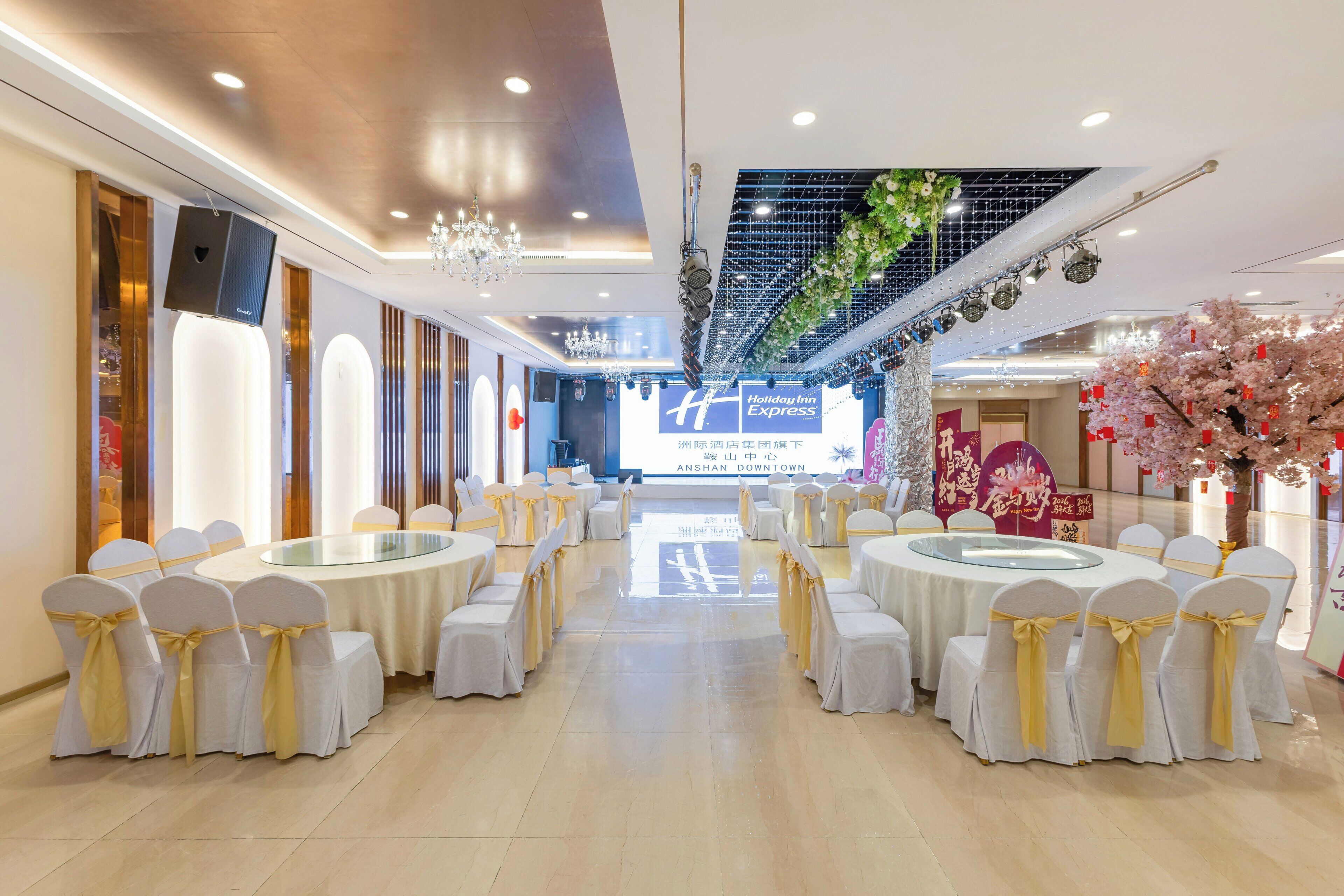 A banquet hall with round tables and chairs, a stage with a large screen, and decorative elements like chandeliers and floral arrangements.