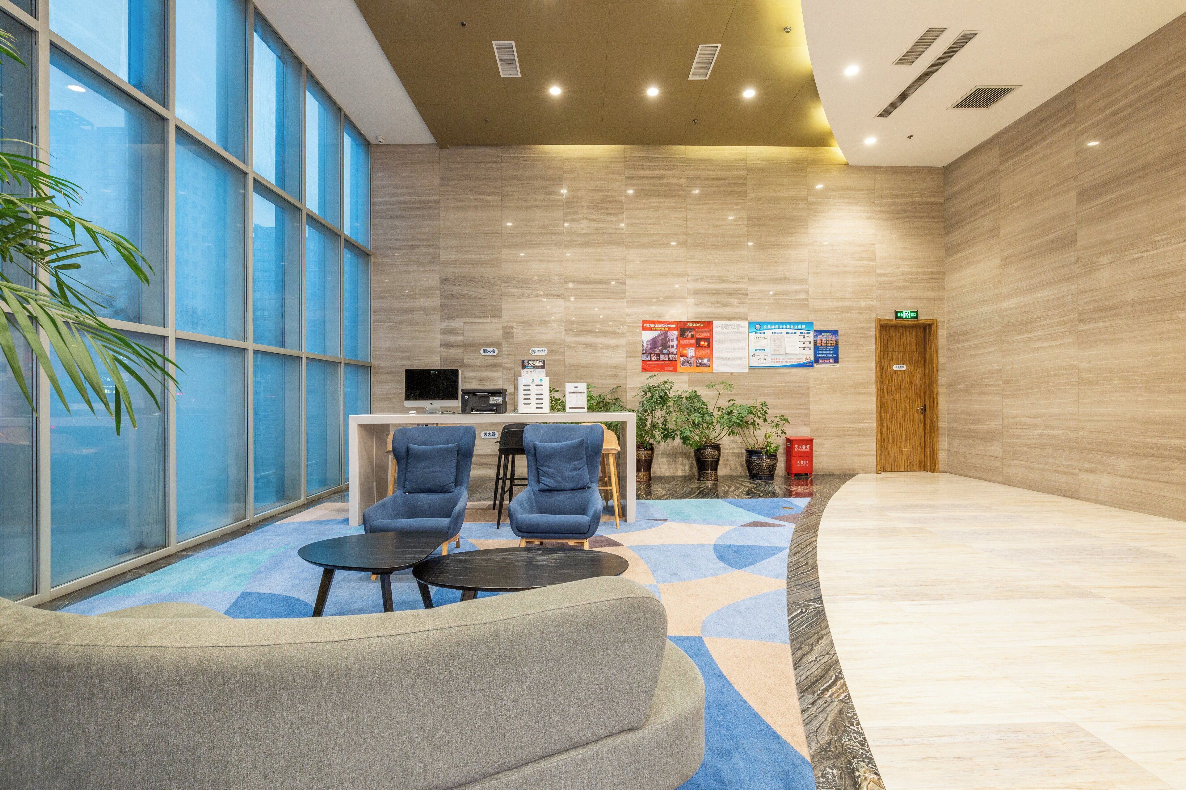 A modern lobby with a reception desk, two blue armchairs, a coffee table, and a large window with a view.
