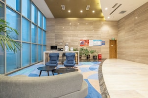 A modern lobby with a reception desk, two blue armchairs, a coffee table, and a large window with a view.