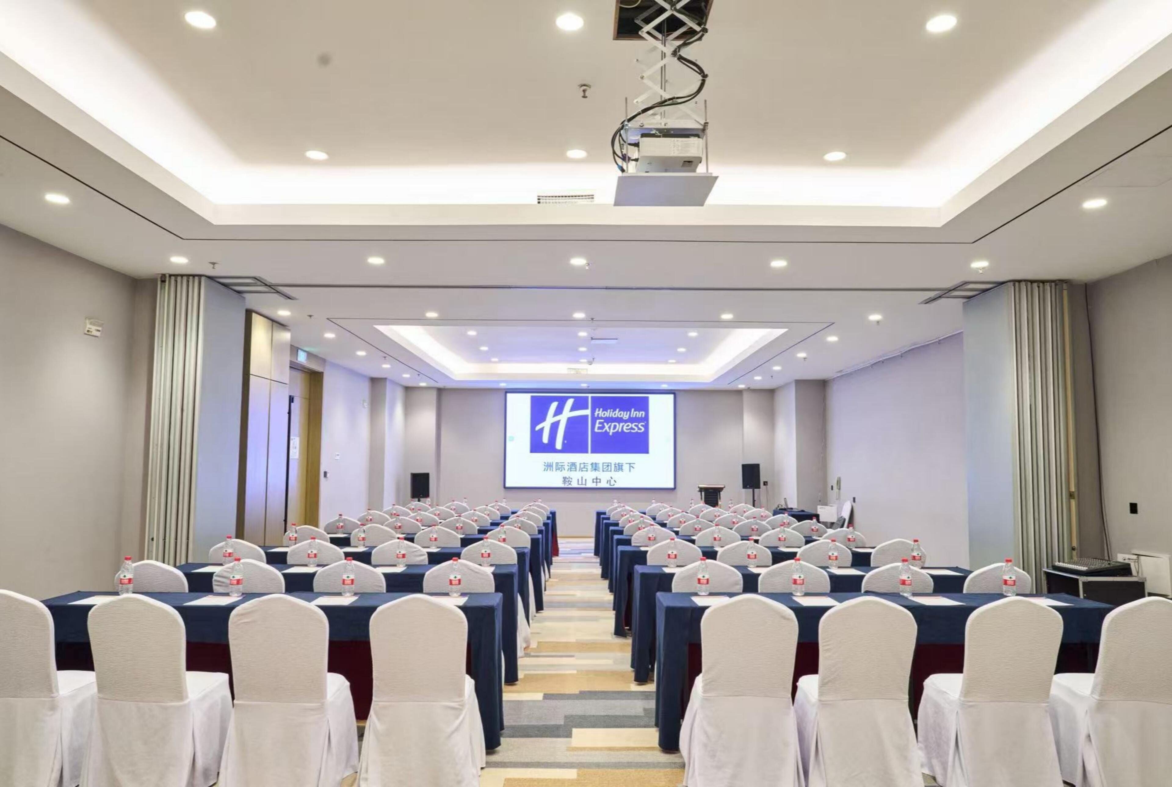 A conference room with rows of tables and chairs, a projector screen, and a sign for Holiday Inn Express.