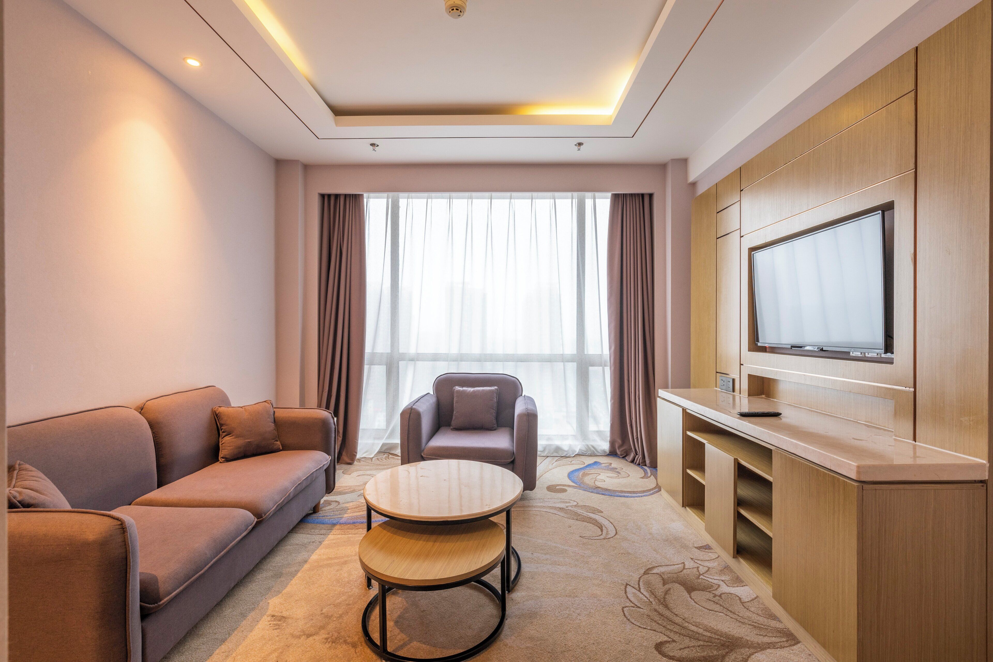A hotel room with a sofa, armchair, round table, and a TV mounted on a wooden cabinet.