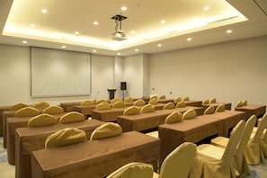 A conference room with rows of tables and chairs, a projector screen, and a speaker.