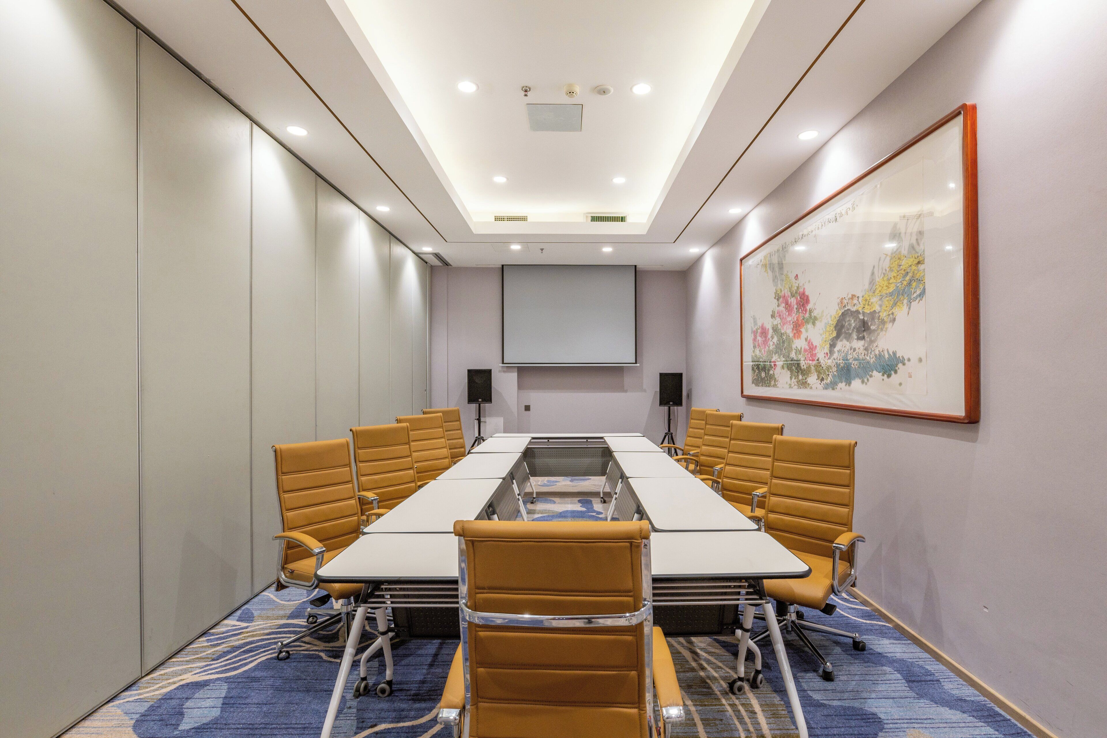 A conference room with a long table, chairs, a projector screen, and a large framed artwork on the wall.