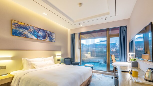 A modern hotel room with a large bed, a desk with a kettle, a chair, and a view of the city through the glass door. - Holiday Inn Express Anshan Downtown by IHG (Anshan)