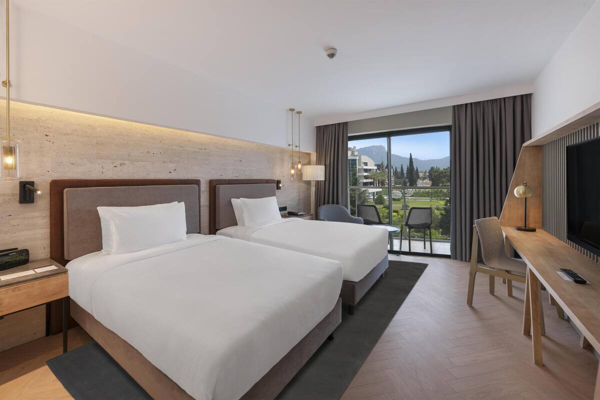 A hotel room with two beds, a desk, a TV, and a balcony with a view.