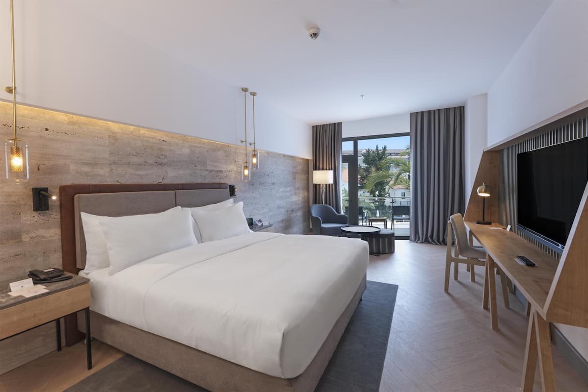 A modern hotel room with a large bed, a desk with a TV, a small table, and a view of the outdoors.