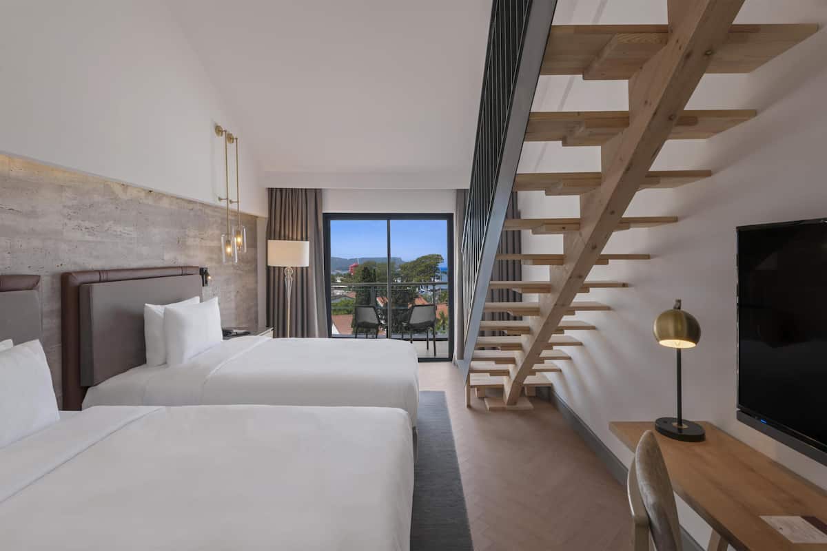 A hotel room with two beds, a TV, a desk, a lamp, and a view of the outdoors.