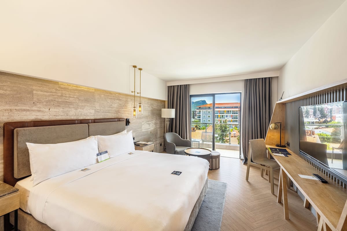 A hotel room with a large bed, a desk, a chair, a lamp, and a TV.