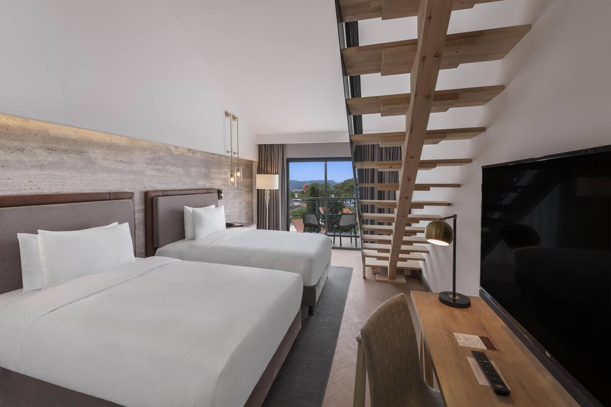 A hotel room with two beds, a TV, a desk, and a balcony with a view.