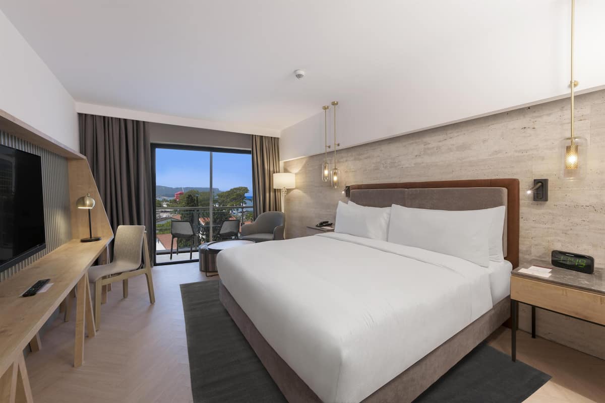 A modern hotel room with a large bed, a desk, a chair, a TV, a balcony with a view, and a clock on the nightstand.
