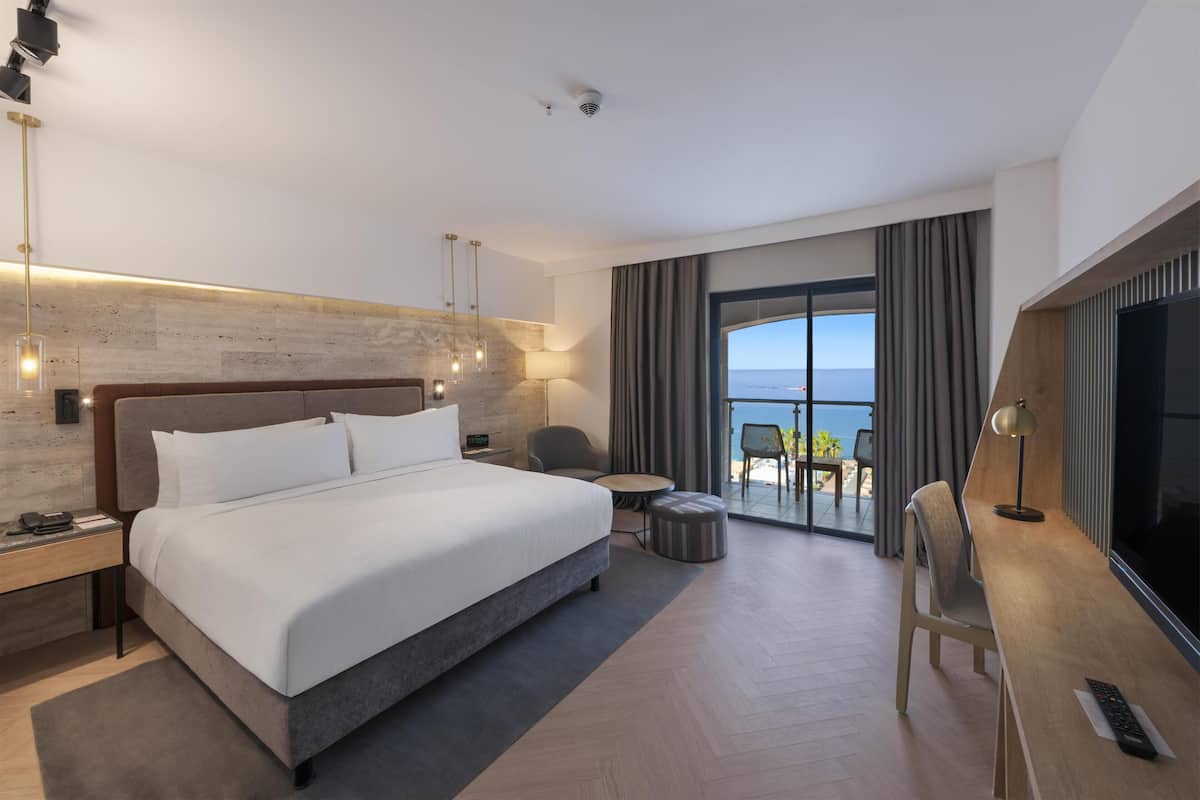 A modern hotel room with a large bed, a desk, a TV, and a balcony with a view of the sea.