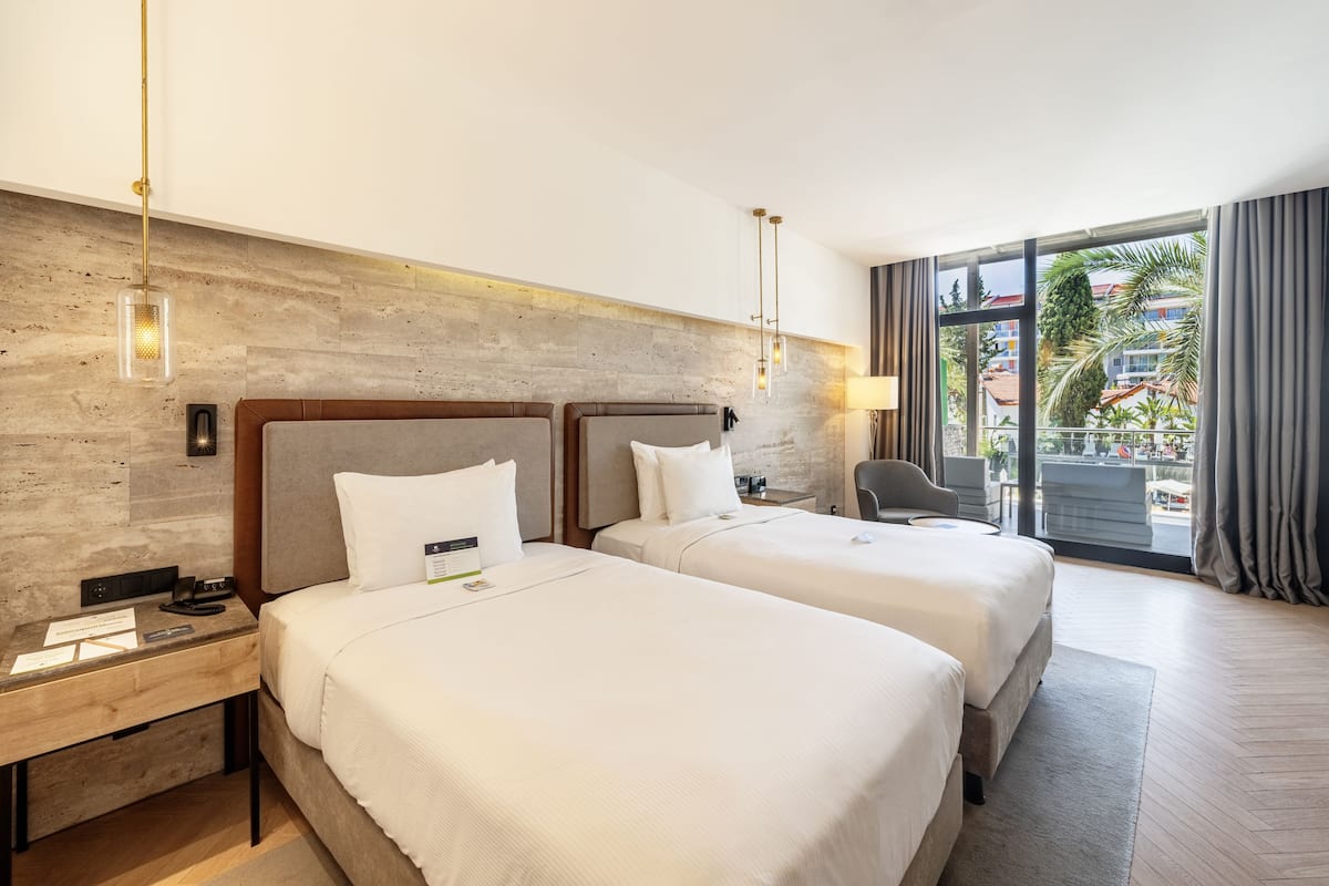 A hotel room with two beds, a wooden headboard, a desk with a lamp, and a view of the outside.