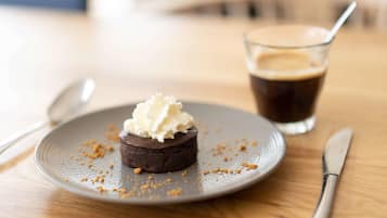 Ein Dessert auf einem Teller, umgeben von Krümeln, ein Glas dunkle Flüssigkeit mit Strohhalm und Besteck auf einem Holztisch.