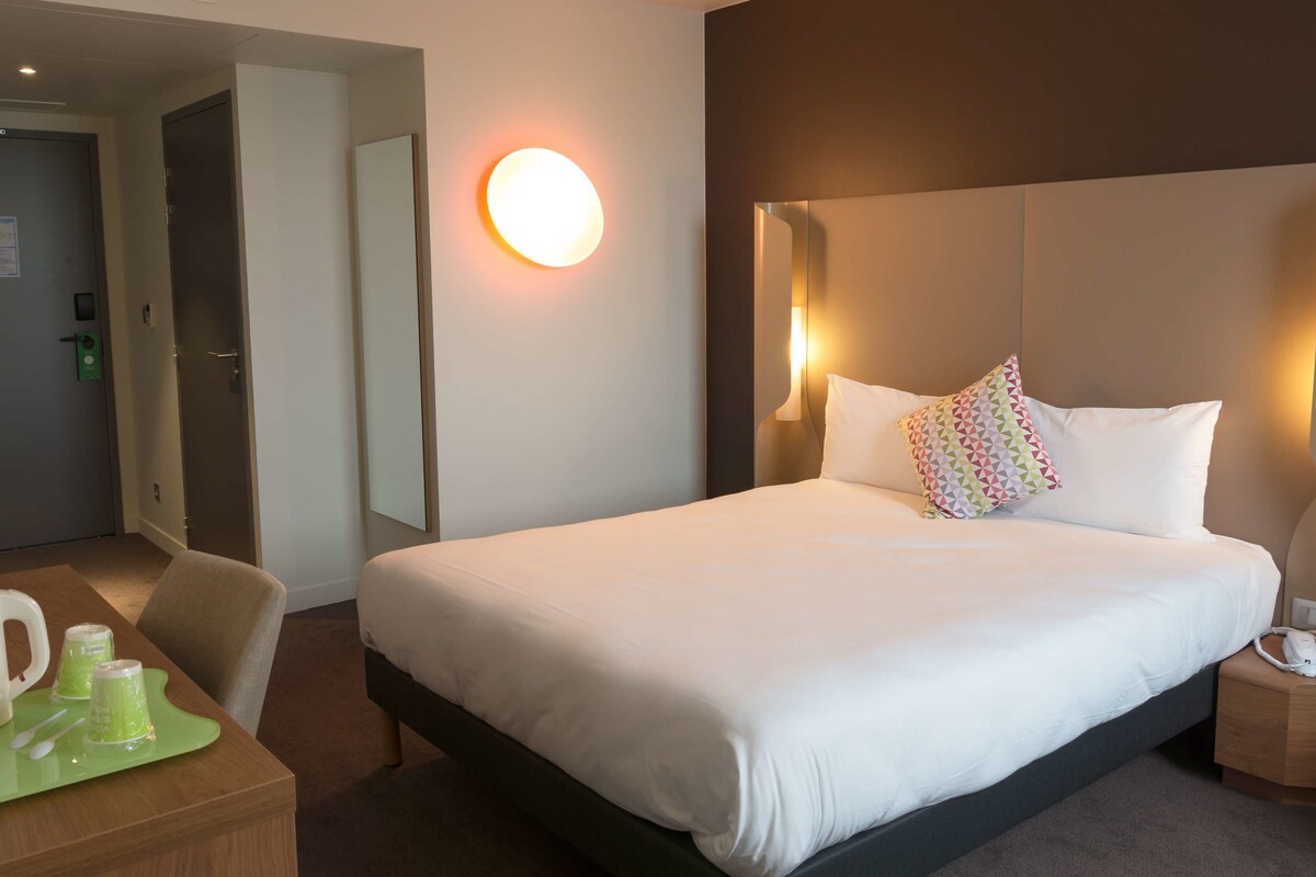 Une chambre d’hôtel avec un grand lit, une table de chevet avec une lampe, une chaise et un bureau équipé d’une machine à café et de verres.