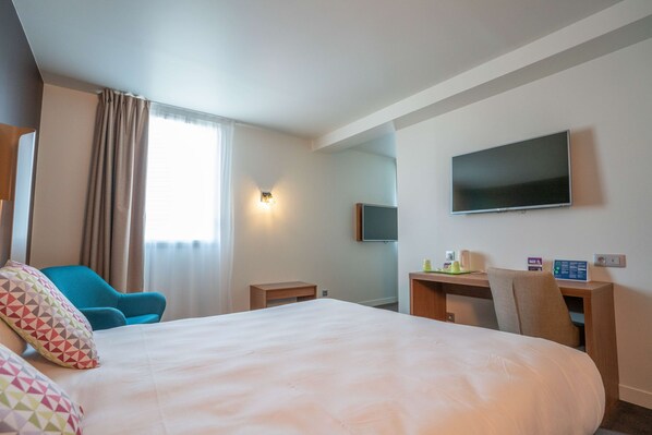 A hotel room with a large bed, a desk with a flat-screen TV, a chair, and a small table.