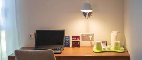 Habitación de hotel con un escritorio de madera, una computadora portátil, una lámpara y una bandeja verde con objetos encima.