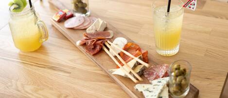 Una mesa de madera con una tabla de fiambres, dos bebidas con pajitas y un vaso de aceitunas.