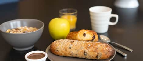 A breakfast setting with a croissant, a bowl of cereal, a small cup of chocolate spread, a glass of orange juice, and a white mug on a dark table.