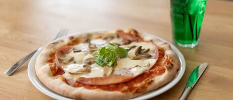 A pizza with ham and mushrooms on a white plate, accompanied by a green drink and cutlery on a wooden table.