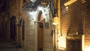 Una calle estrecha con la entrada de un hotel, un cartel sobre la puerta y un muro de ladrillo a la derecha.