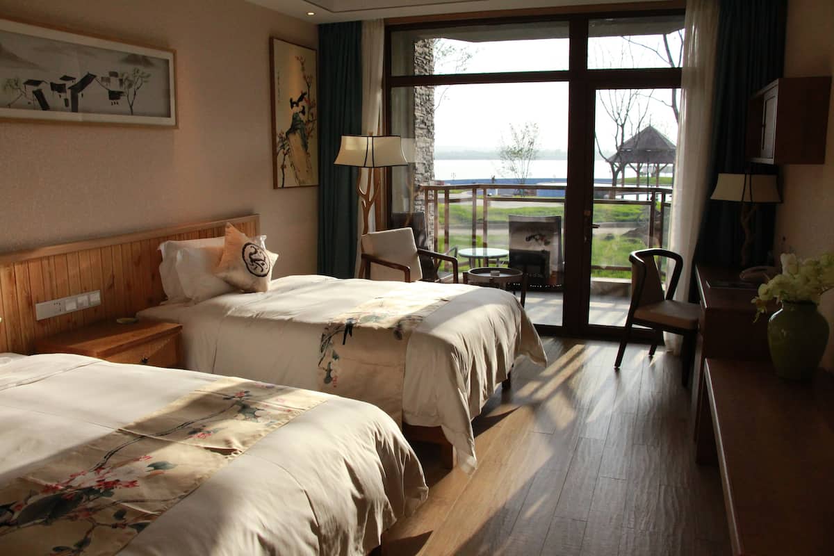 Habitación de hotel con dos camas, cabecera de madera, mesita de noche, escritorio con silla, lámpara y vista a un lago y un pabellón a través de grandes ventanales.