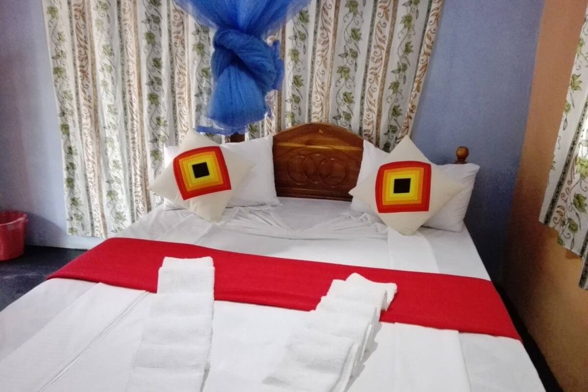 A neatly made bed with white linens and a red bedspread, two pillows with a yellow and red design, and a blue curtain.