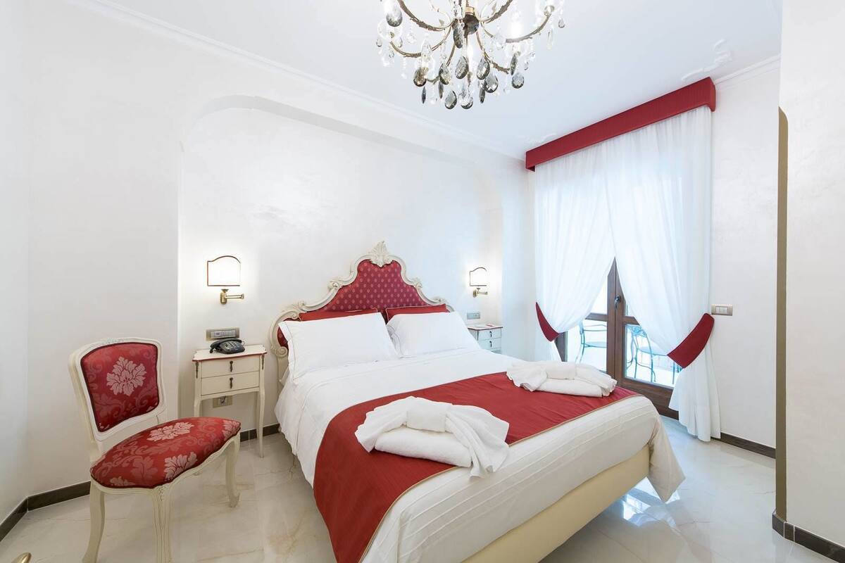 Une chambre d’hôtel avec un grand lit, une chaise, une table de chevet, un lustre et une fenêtre avec des rideaux.