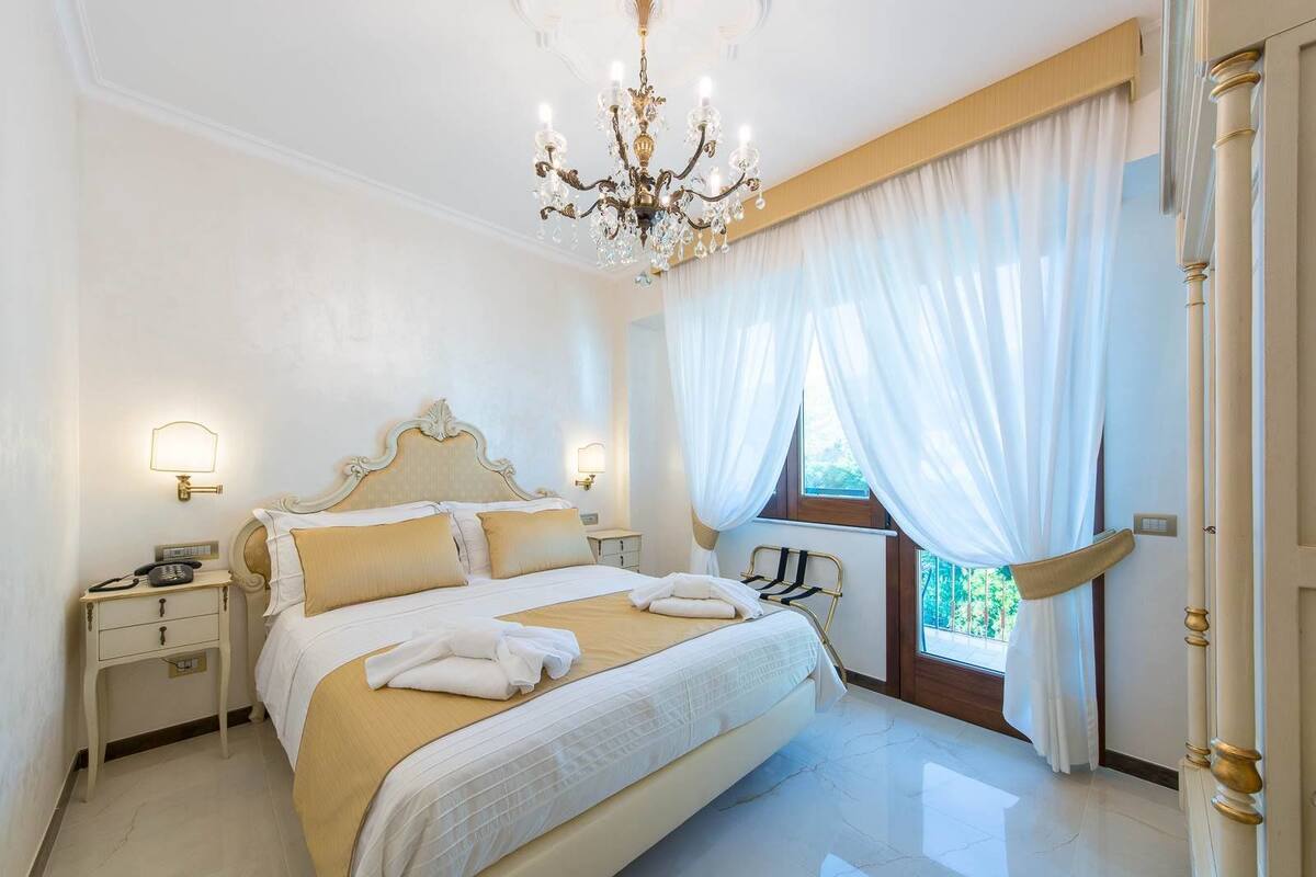Une chambre spacieuse avec un grand lit, des tables de chevet, un lustre et une vue sur la verdure par la fenêtre.