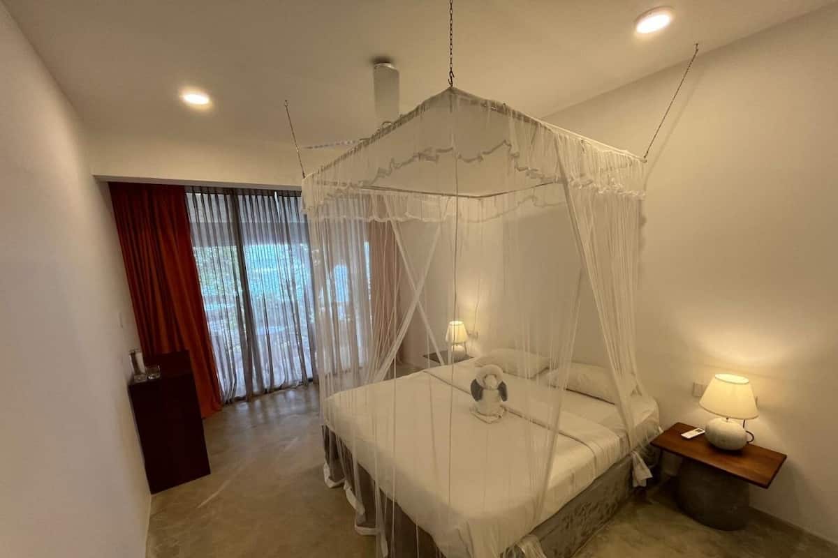 A bedroom with a four-poster canopy bed, a bedside table with a lamp, and a view of a balcony with curtains.