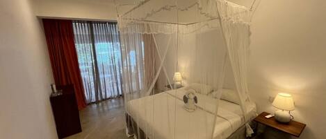 A bedroom with a four-poster canopy bed, a bedside table with a lamp, and a view of a balcony with curtains.
