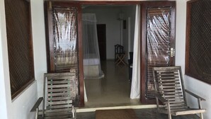 Une porte d’entrée avec deux chaises en bois devant, un paillasson et, à travers la porte, on aperçoit une pièce avec un lit et une chaise.