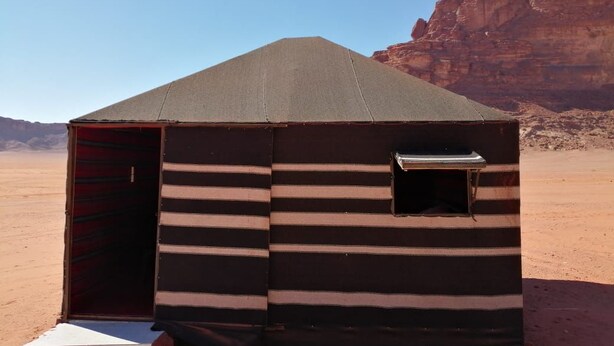 Uma pequena construção com listras, um cômodo único, com porta de metal e pequena janela, emoldurada por uma paisagem desértica com formações rochosas avermelhadas.