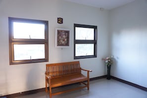 Una habitación con dos ventanas, un banco de madera, un cuadro enmarco y un jarrón con flores.